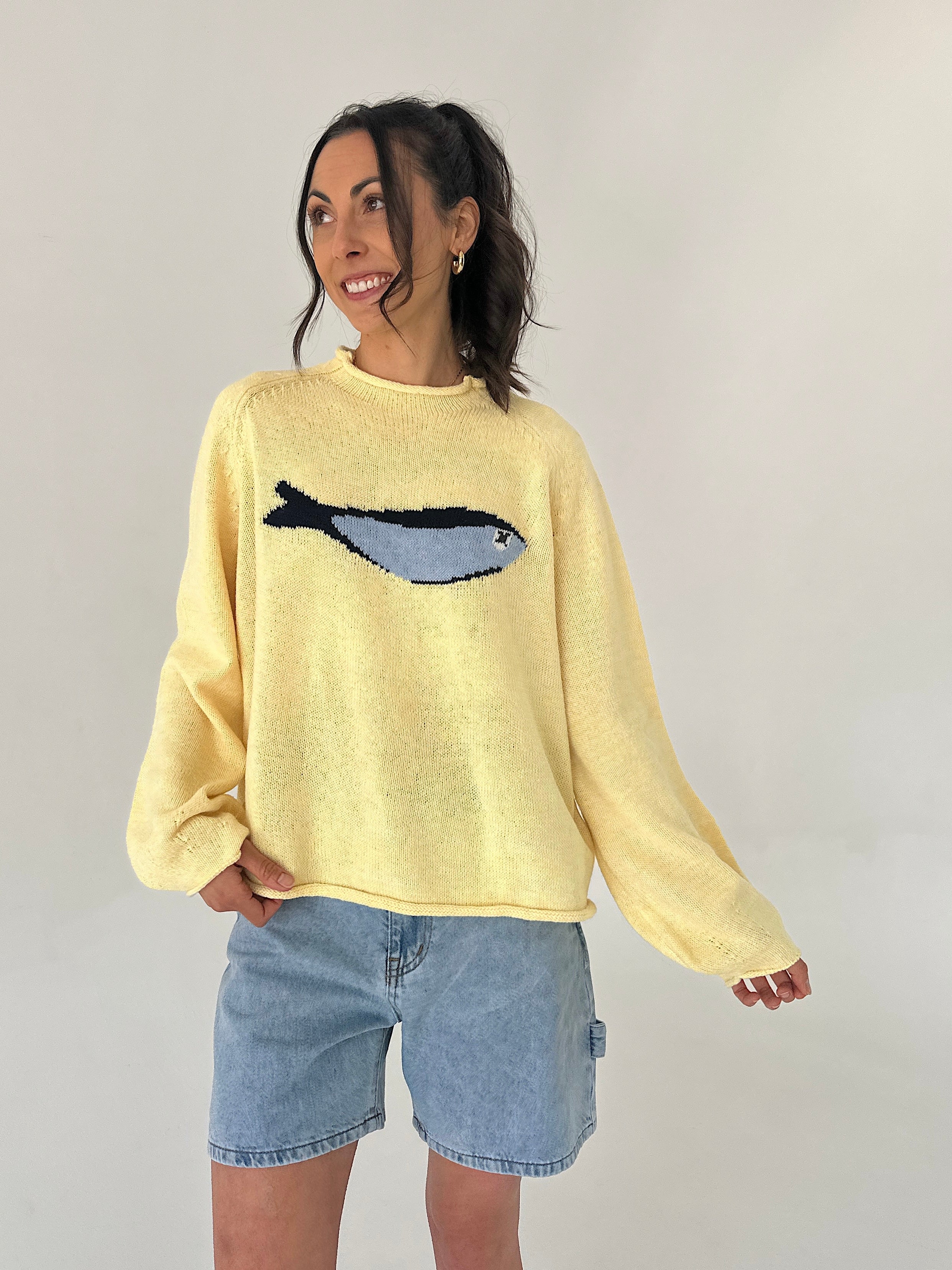 Woman wearing a yellow sweater with a fish design, standing against a plain background