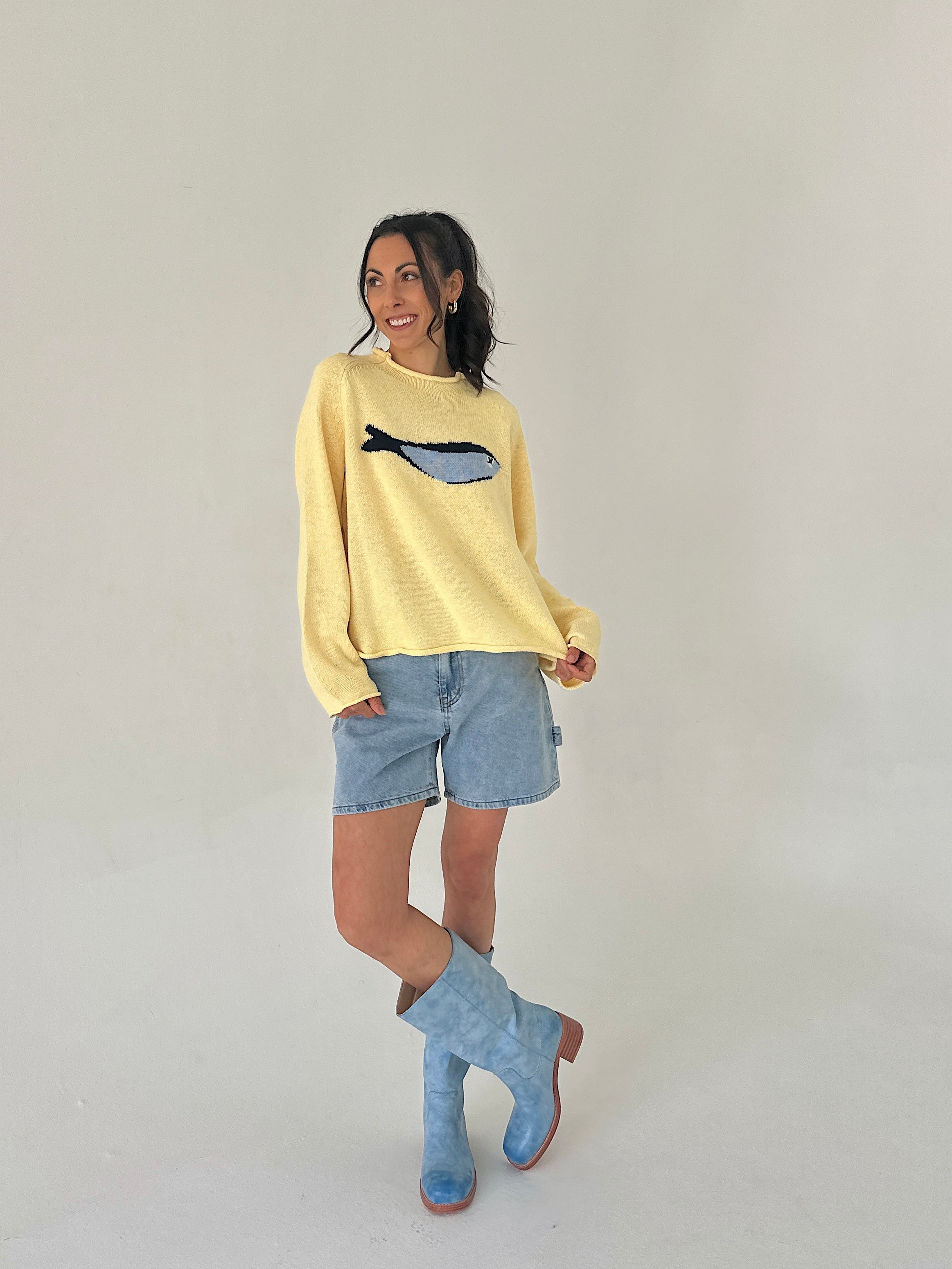 Woman wearing a yellow sweater with a fish design, denim shorts, and blue boots on a plain background