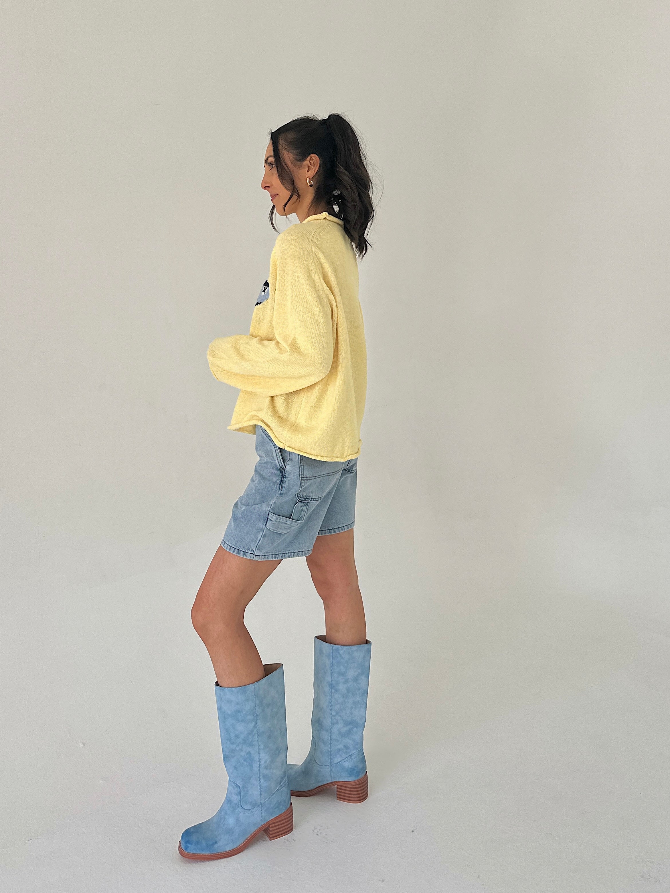 Person wearing a yellow sweater, denim shorts, and blue boots on a plain background