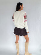 Person wearing a cream sweater with patterned sleeves, dark pleated skirt, and beige knee-high boots on a plain background