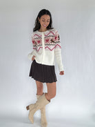 Woman wearing a patterned cardigan, skirt, and knee-high boots on a plain background