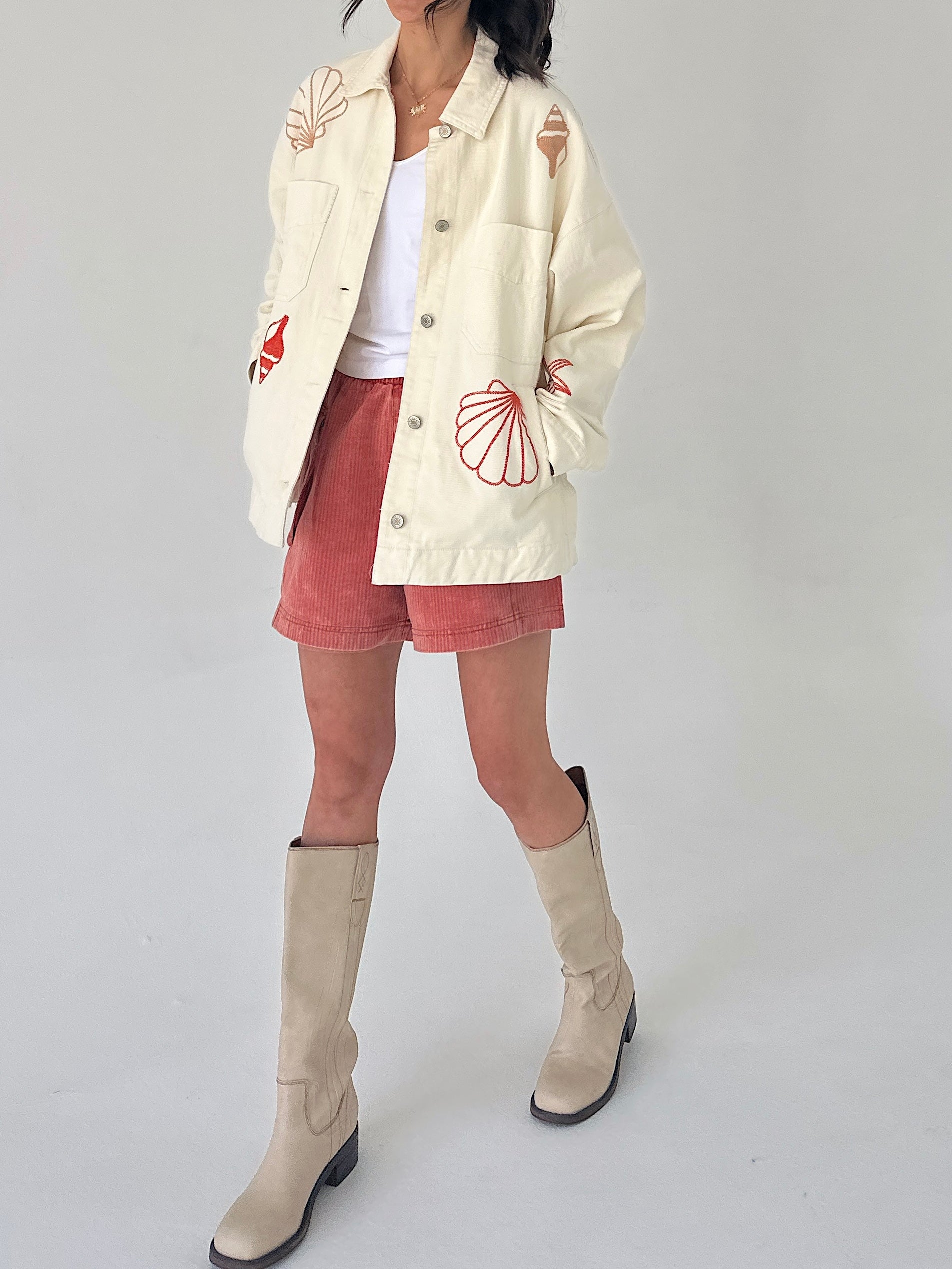 Person wearing a cream jacket with red patterns, rust shorts, and beige knee-high boots on a white background