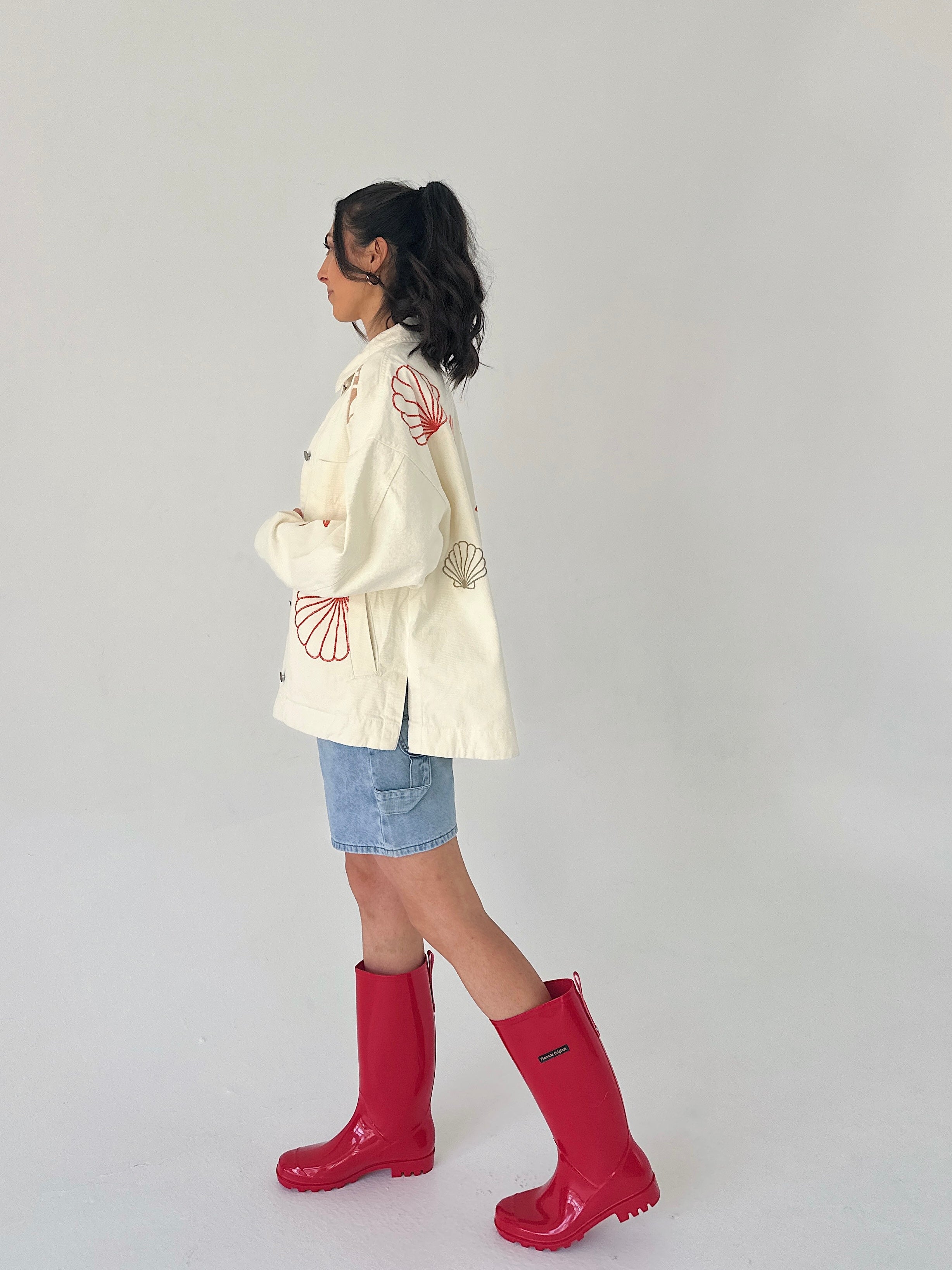 Person wearing a white jacket with red patterns, denim shorts, and red rain boots on a plain background