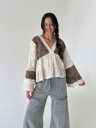 A woman modeling a bohemian-style long-sleeve blouse with cream waffle-knit fabric, lace trim, and brown floral patterned panels, paired with wide-leg gray pants, against a plain off-white background.