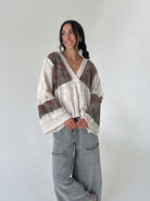 A woman modeling a bohemian-style patchwork blouse with floral and textured panels, lace trim, and wide sleeves, paired with gray wide-leg cargo pants against a plain light gray background.