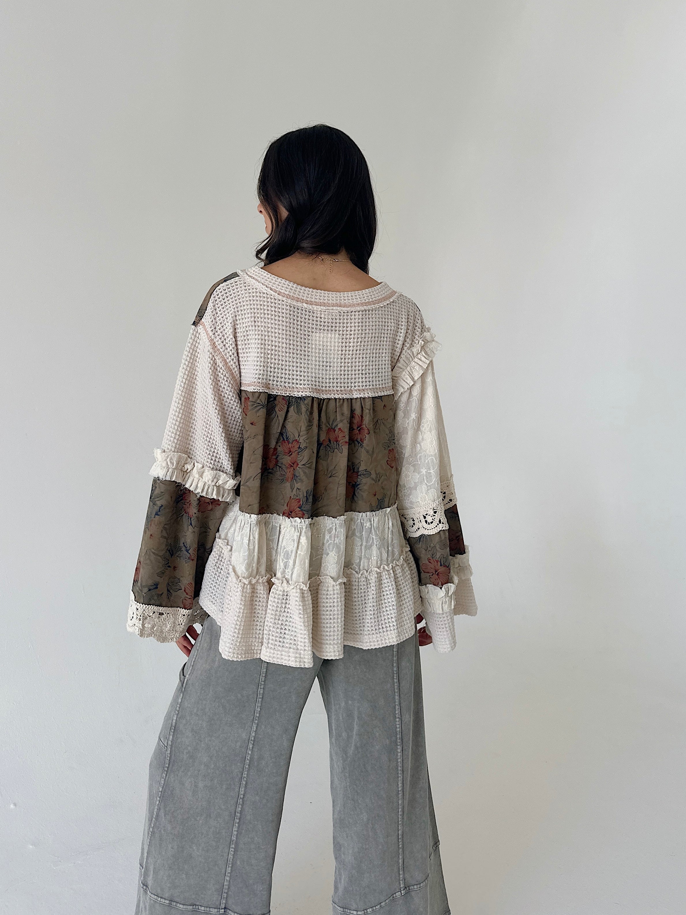 A woman viewed from behind wearing a patchwork long-sleeve blouse with cream waffle knit, floral brown panels, lace trim, and wide-leg gray pants against a plain light gray wall.