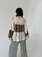 A woman viewed from behind wearing a patchwork long-sleeve blouse with cream waffle knit, floral brown panels, lace trim, and wide-leg gray pants against a plain light gray wall.