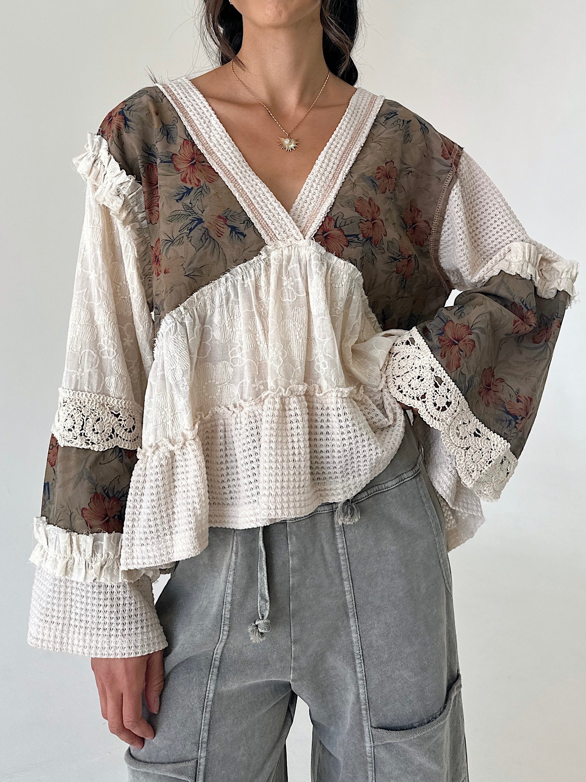 A woman wearing a bohemian-style blouse with a mix of floral printed mesh and textured cream lace, paired with gray drawstring pants.