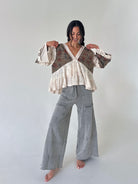 A woman modeling a bohemian-style patchwork top with wide sleeves, lace trim, and a tiered ruffle hem, paired with wide-leg gray pants, against a plain light gray studio background.