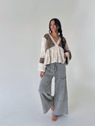 A woman modeling a bohemian-style outfit consisting of a cream and brown patterned long-sleeve blouse with lace trim and wide-leg gray linen pants, standing against a plain light gray studio background.