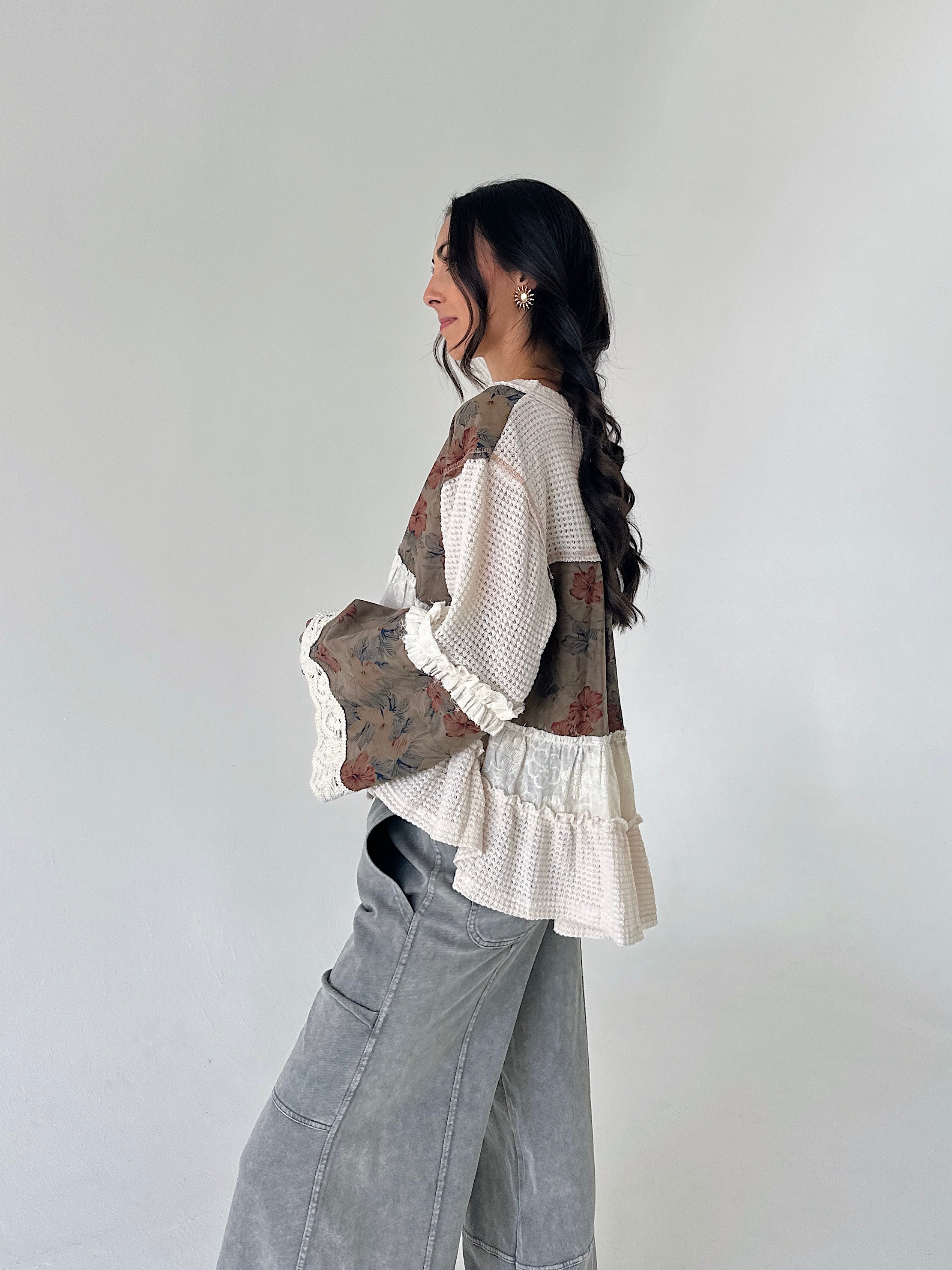 A woman wearing a bohemian-style patchwork blouse with cream waffle knit, floral printed panels, and ruffle trim, paired with wide-leg gray jeans, standing against a plain light gray wall.
