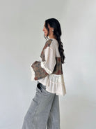 A woman wearing a bohemian-style patchwork blouse with cream waffle knit, floral printed panels, and ruffle trim, paired with wide-leg gray jeans, standing against a plain light gray wall.