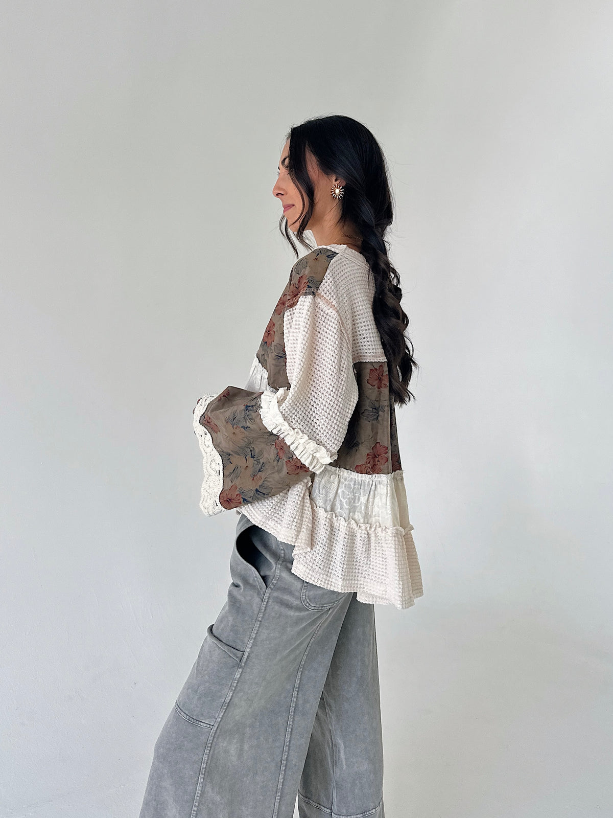 A woman wearing a bohemian-style patchwork blouse with cream waffle knit, floral printed panels, and ruffle trim, paired with wide-leg gray jeans, standing against a plain light gray wall.