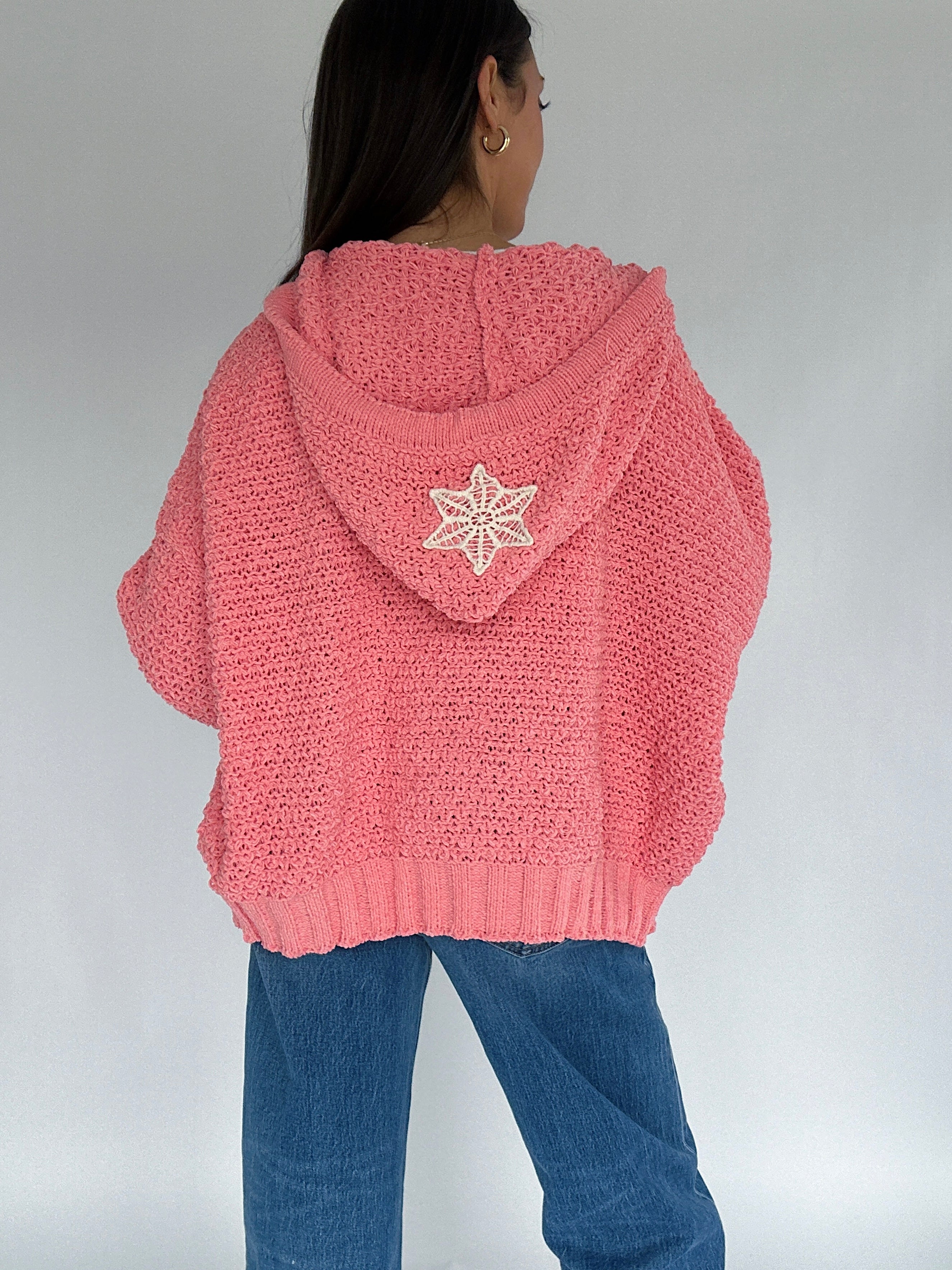 Person wearing a pink knitted hoodie with a crochet patch on the back against a plain background
