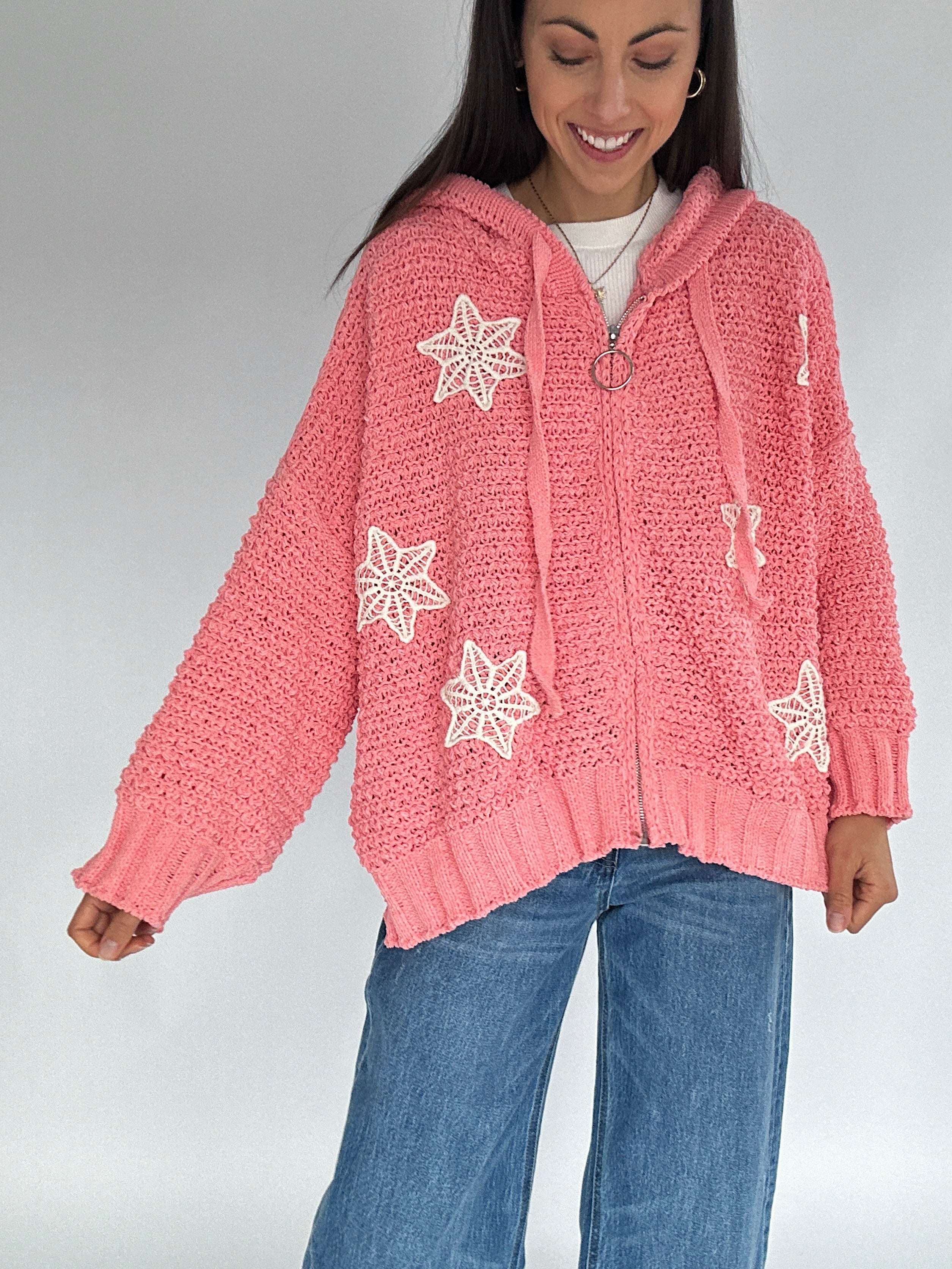 Woman wearing a pink knitted zip up cardigan with crocket patterns on a plain background