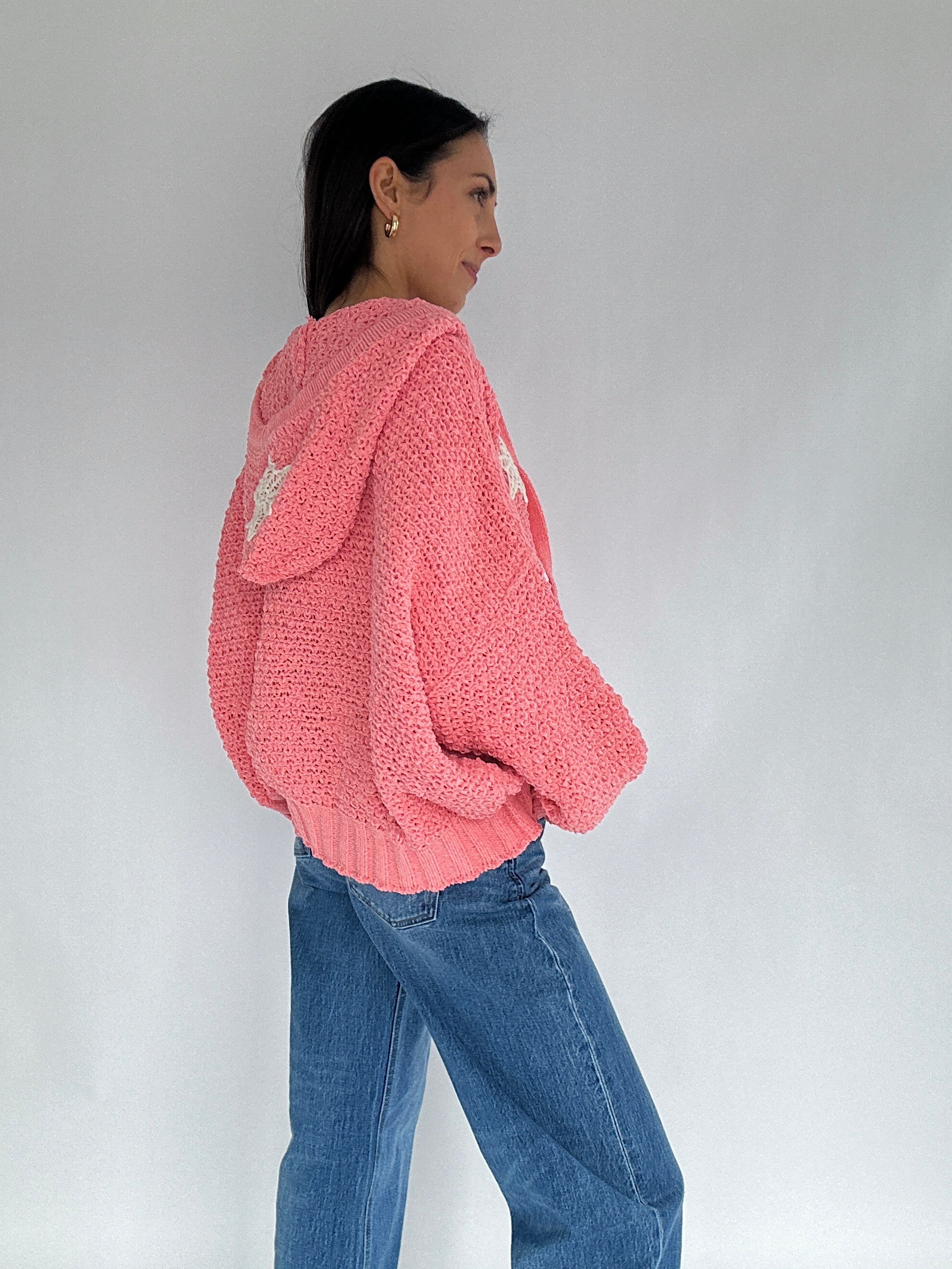 Person wearing a pink knitted hoodie sweater and blue jeans against a plain background