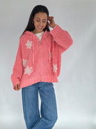 Woman wearing a pink knitted zip up hoodie sweater with white crochet patterns on a plain background