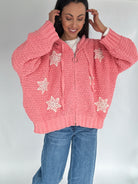 Woman wearing a pink knitted hoodie zip up cardigan with crochet patterns on a plain background