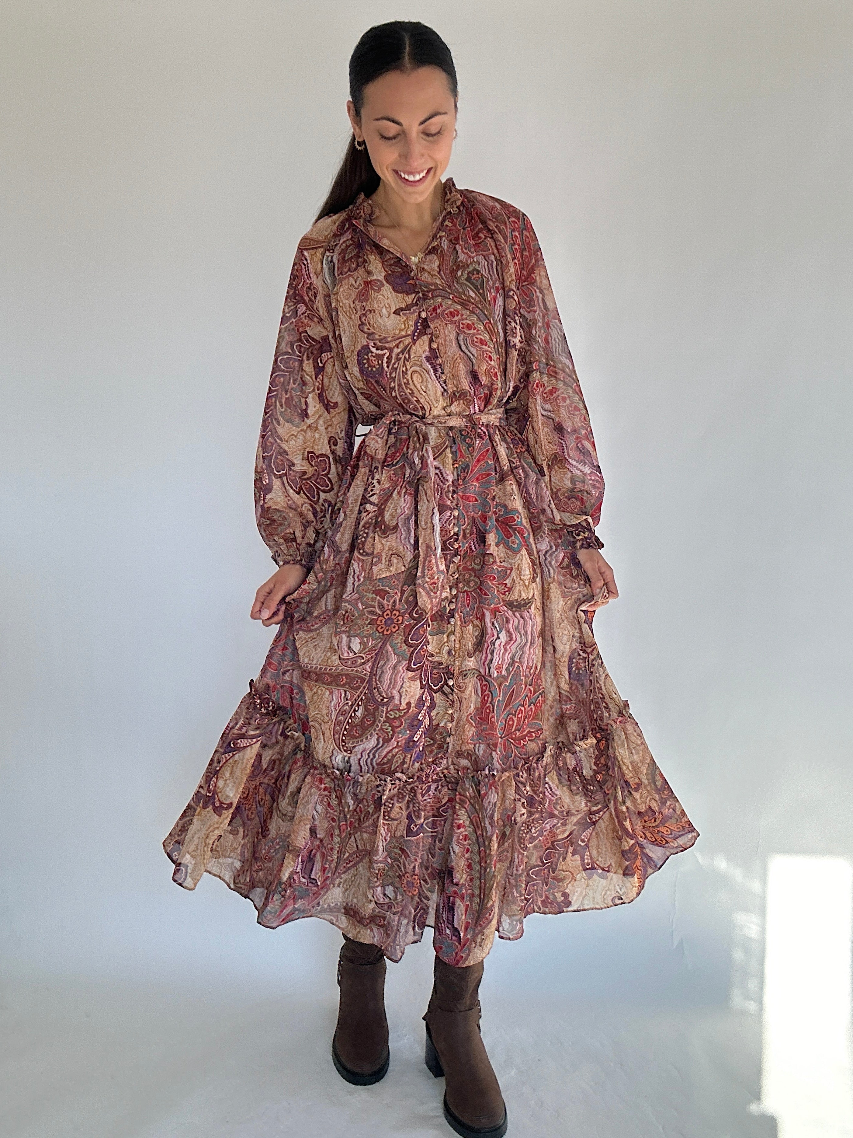 Woman wearing a long, boho paisley patterned dress with a ruffled bottom and waist tie against a white background