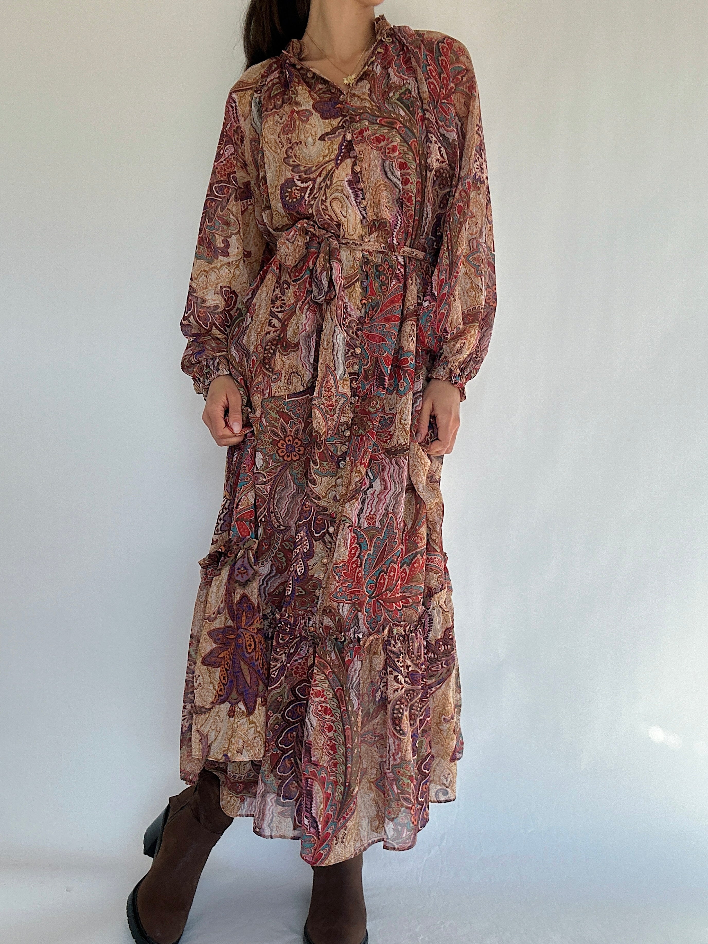 Long boho paisley patterned long sleeve dress with button front, waist tie and ruffled bottom worn by a person on a white background