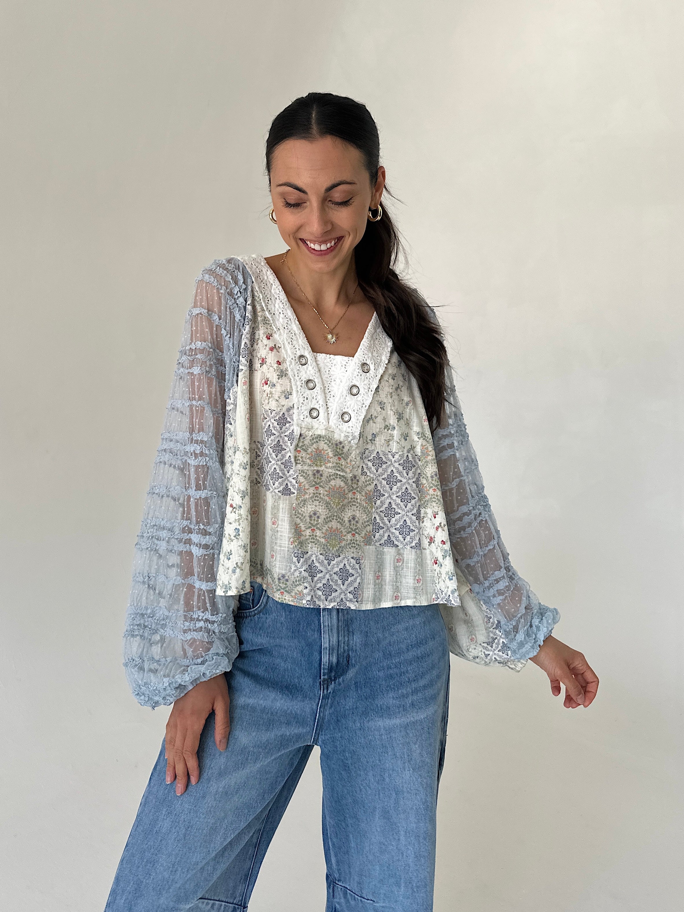 A woman modeling a printed blouse with sheer light blue patterned long sleeves, paired with blue jeans, against a plain light gray studio background.