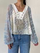 A woman wearing a bohemian-style patchwork blouse with sheer light blue lace long sleeves and a cream-colored embroidered bodice, paired with blue jeans.