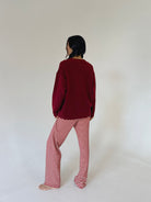 A person standing barefoot against a plain off-white studio background, viewed from the back. They are wearing a dark burgundy long-sleeve top and red-and-white horizontally striped pants.