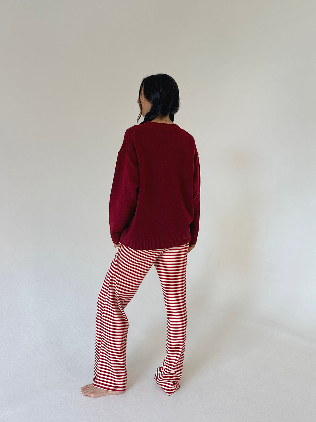 A person standing barefoot against a plain off-white studio background, viewed from the back. They are wearing a dark burgundy long-sleeve top and red-and-white horizontally striped pants.