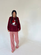 A woman modeling a cozy holiday outfit consisting of a deep burgundy long-sleeve top with a red goose graphic and matching red-and-white striped pants, standing against a plain off-white studio background.
