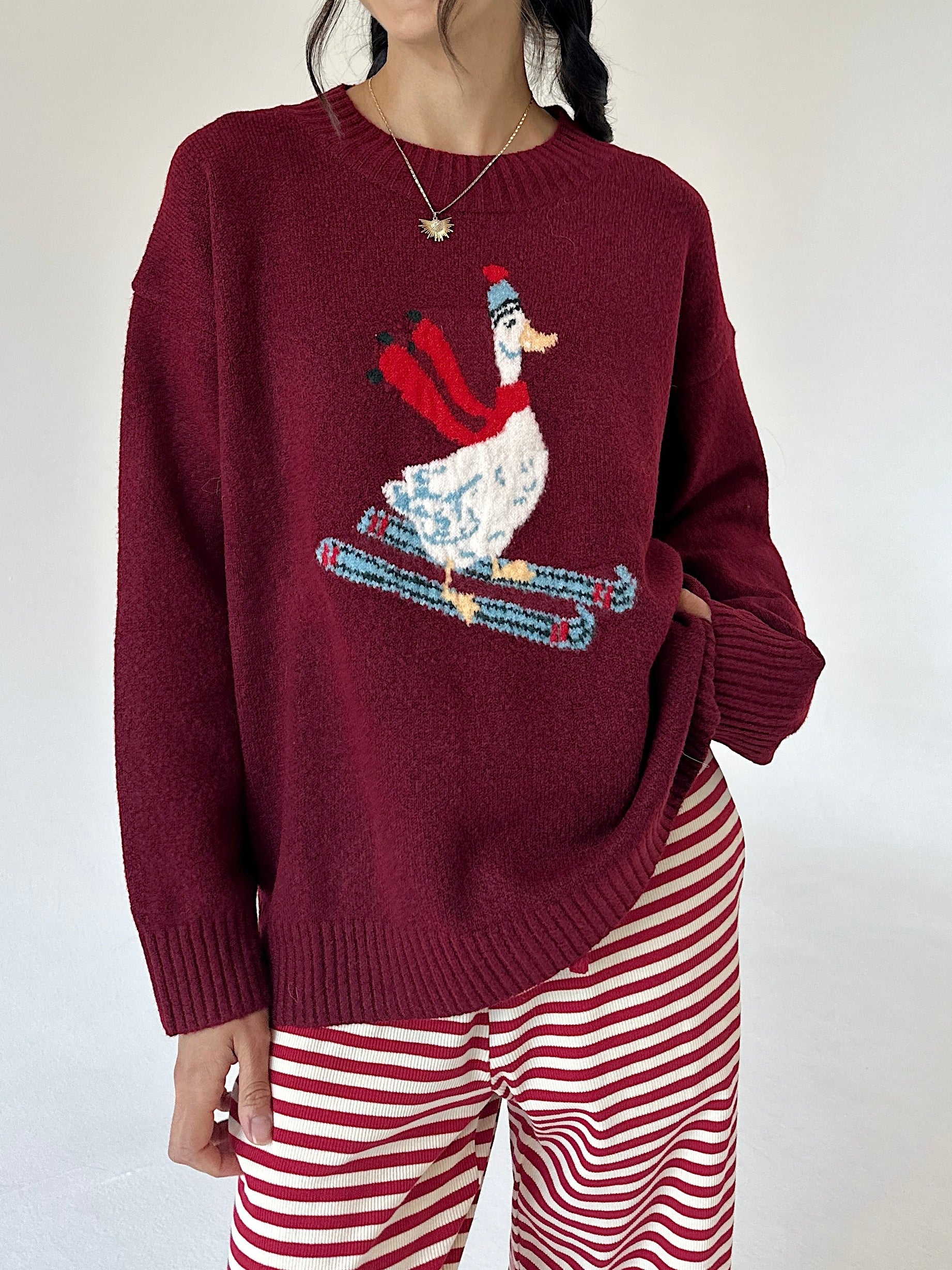 A person wearing a burgundy knit sweater with a large embroidered skiing duck design on the front, paired with red and white striped pants, against a plain light background.