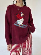 A person wearing a burgundy knit sweater with a large embroidered skiing duck design on the front, paired with red and white striped pants, against a plain light background.
