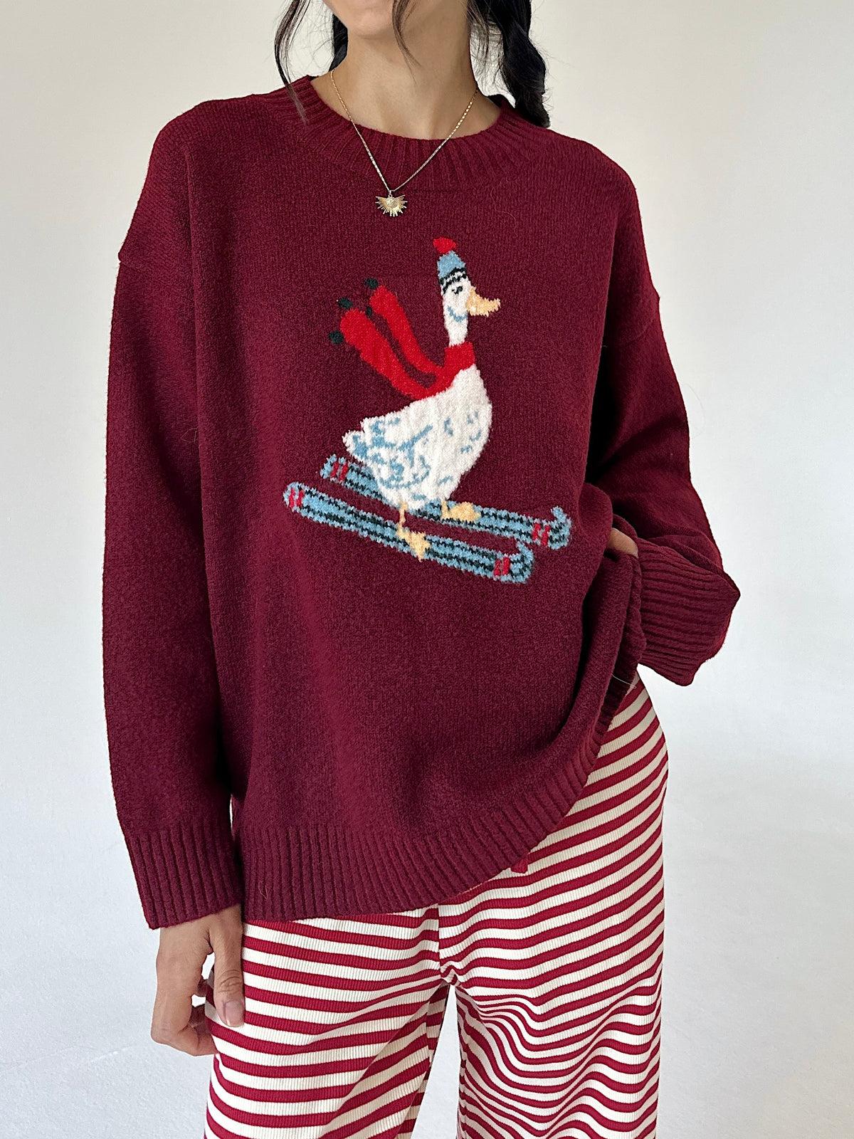 A person wearing a burgundy knit sweater with a large embroidered skiing duck design on the front, paired with red and white striped pants, against a plain light background.