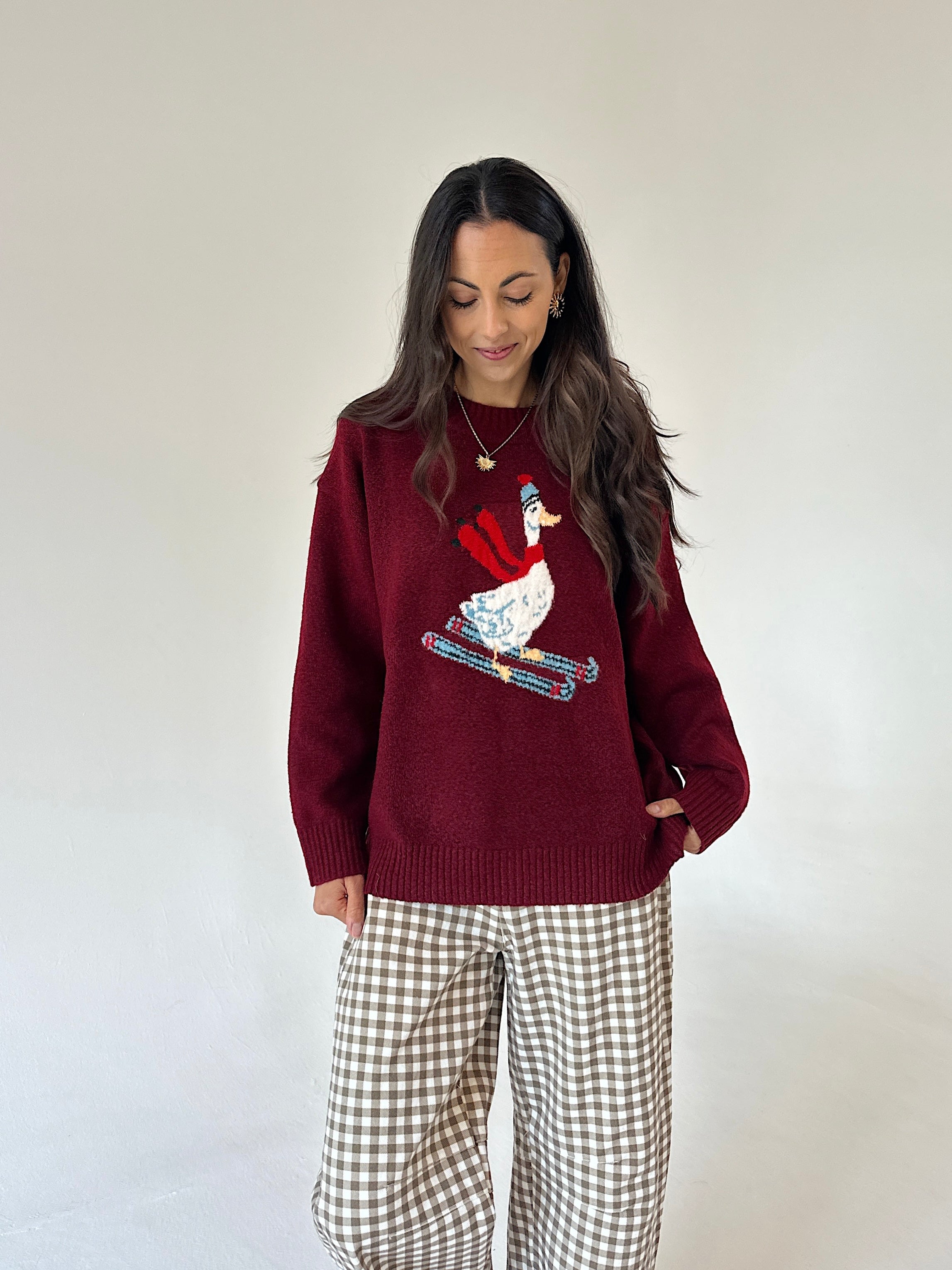 A woman wearing a burgundy knit sweater with a colorful embroidered skiing goose design on the front, paired with brown and white gingham wide-leg pants, standing against a plain off-white background.