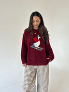 A woman wearing a burgundy knit sweater with a colorful embroidered skiing goose design on the front, paired with brown and white gingham wide-leg pants, standing against a plain off-white background.