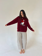 A woman stands barefoot against a plain off-white studio background, wearing a burgundy knit sweater with a white goose graphic on the front and beige-and-white gingham pajama pants.