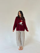 A woman stands barefoot against a plain off-white studio background wearing a burgundy long-sleeve knit top with a white goose graphic on the chest and beige-and-white gingham cropped pants.