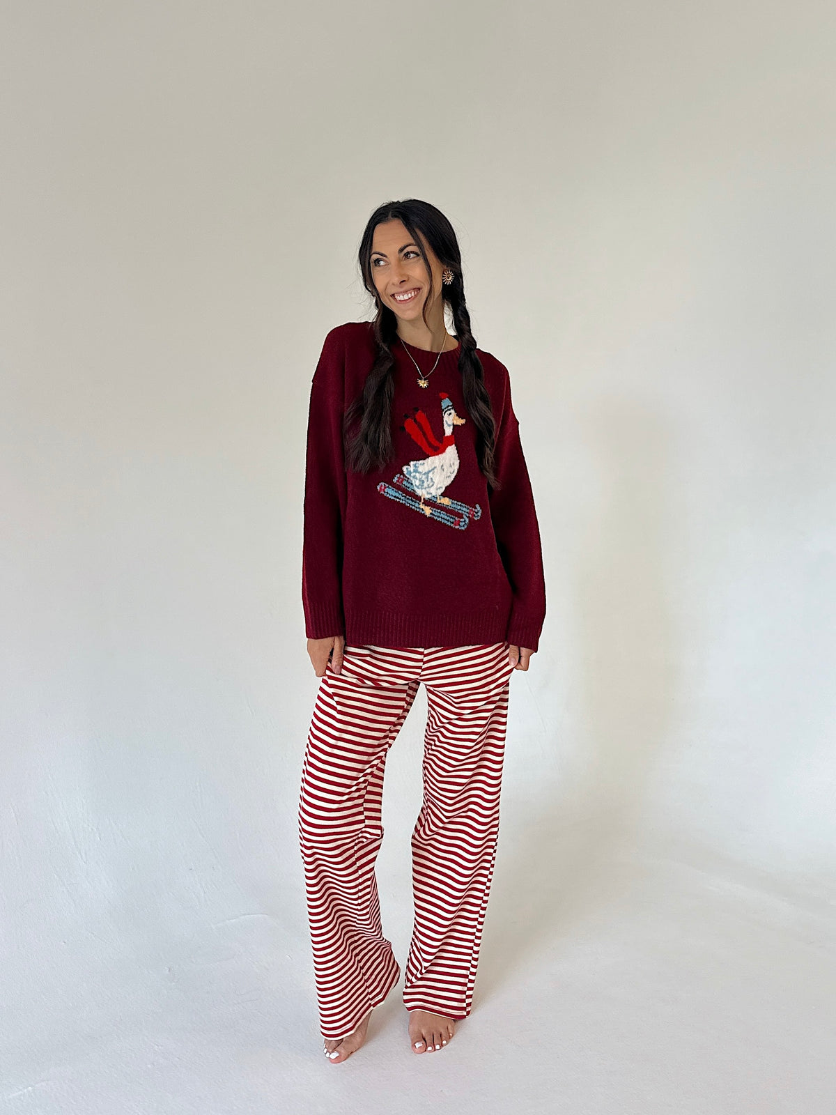 A woman modeling a holiday-themed outfit consisting of a burgundy long-sleeve top with a red, white, and blue goose graphic, paired with red and white horizontally striped pants, against a plain white studio background.