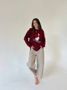A woman stands barefoot against a plain off-white studio background, wearing a burgundy long-sleeve crewneck sweatshirt with a white duck graphic on the front, paired with beige and white gingham wide-leg pants.