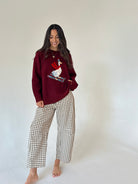 A woman stands barefoot against a plain off-white studio background, wearing a burgundy knit sweater with a white goose graphic on the front and wide-leg beige-and-white gingham pants.