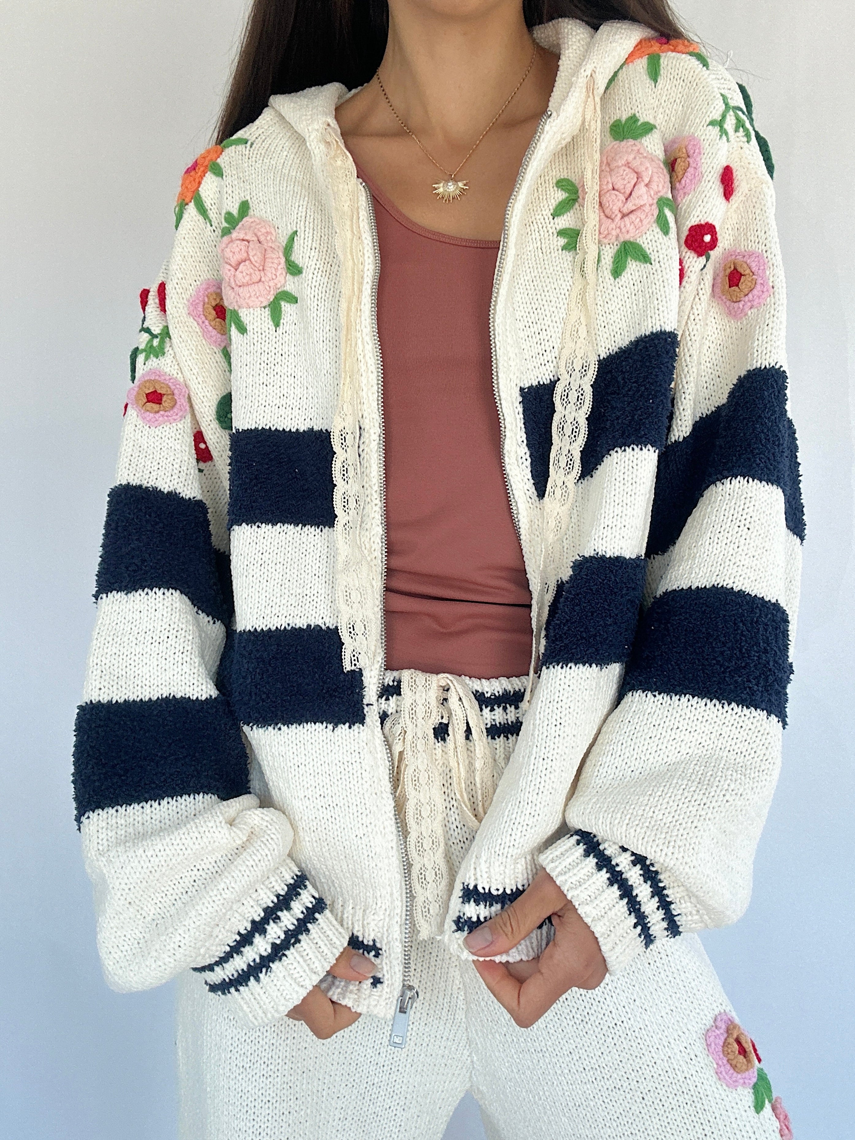 Person wearing a white cardigan with floral patterns and navy blue stripes on a light gray background unzipped
