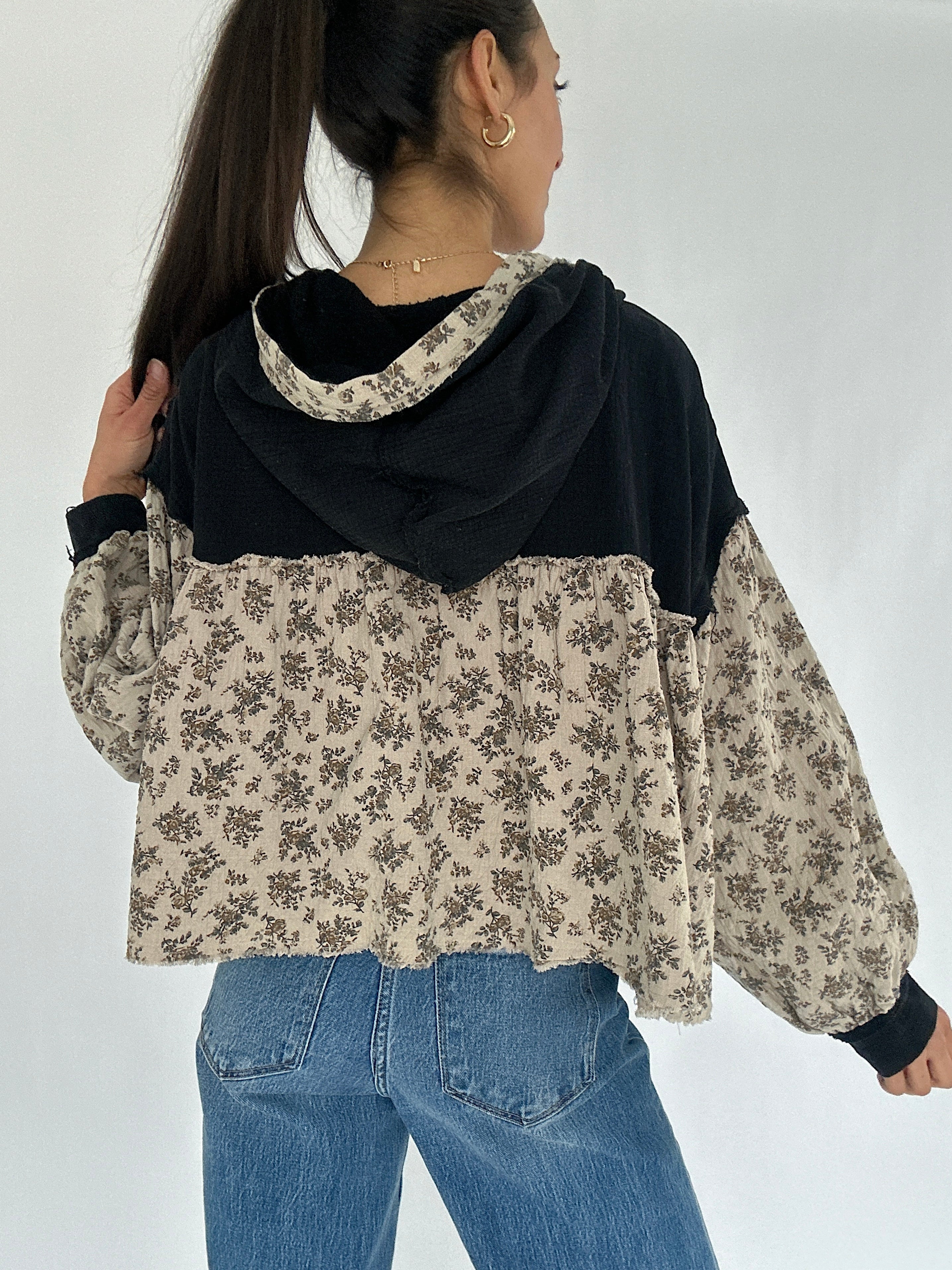 Person wearing a black and beige floral-patterned hoodie top with blue jeans on a white background showing the back