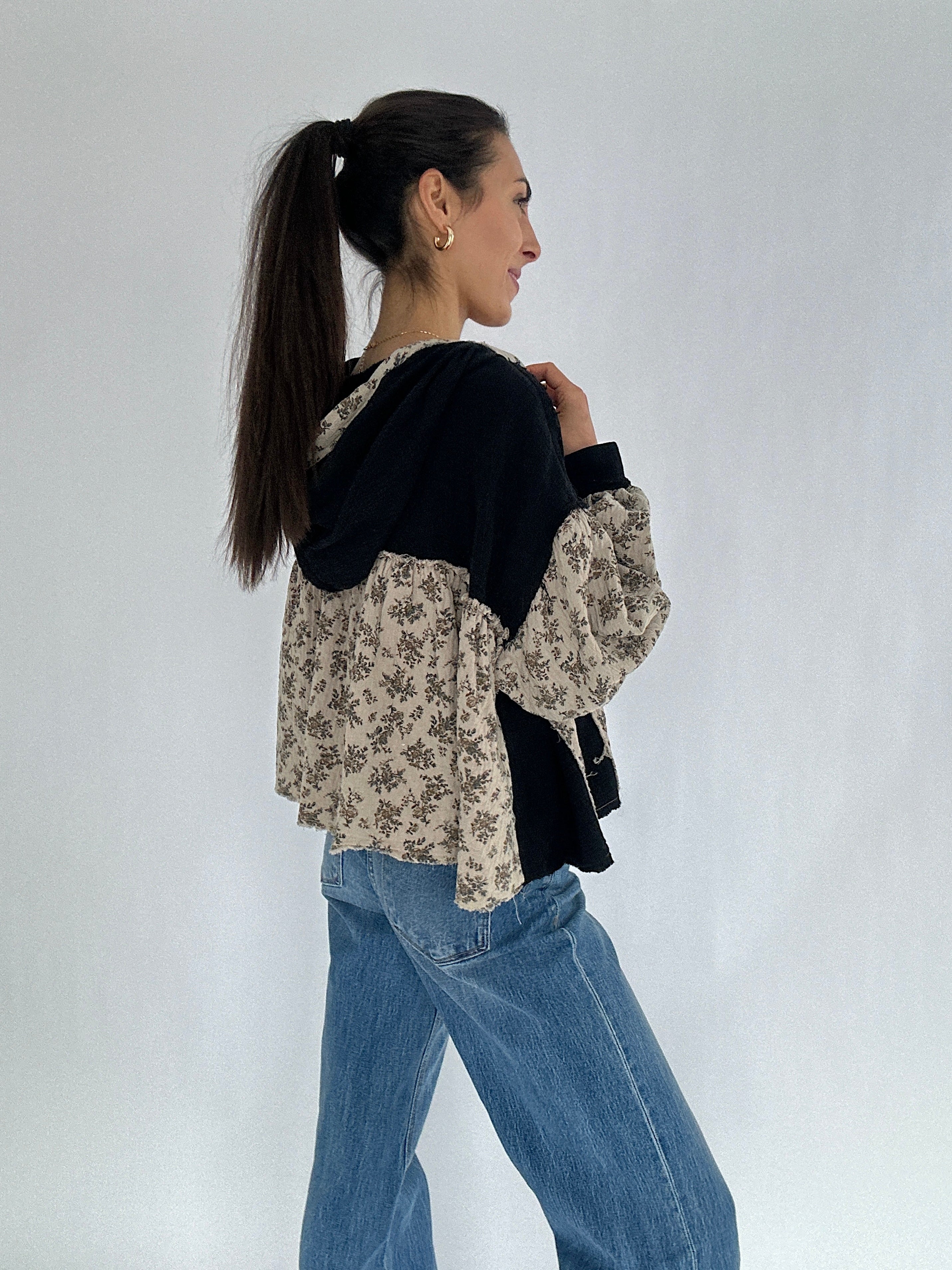 Woman wearing a black and floral hooded top and blue jeans on a white background showing side angle