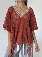 A woman wearing a rust-colored crochet lace top with a V-neck and patterned gray pants, photographed against a plain light background.