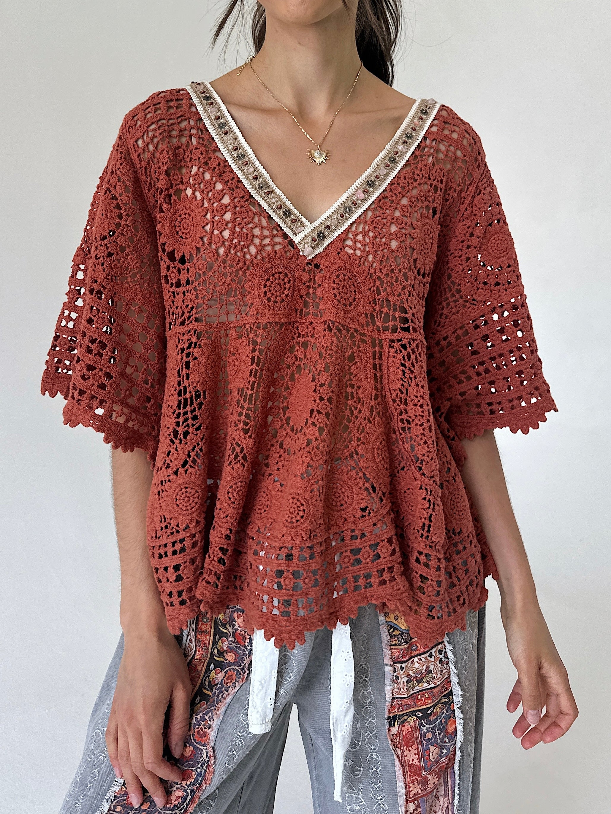 A woman wearing a rust-colored crochet lace top with a V-neck and patterned gray pants, photographed against a plain light background.