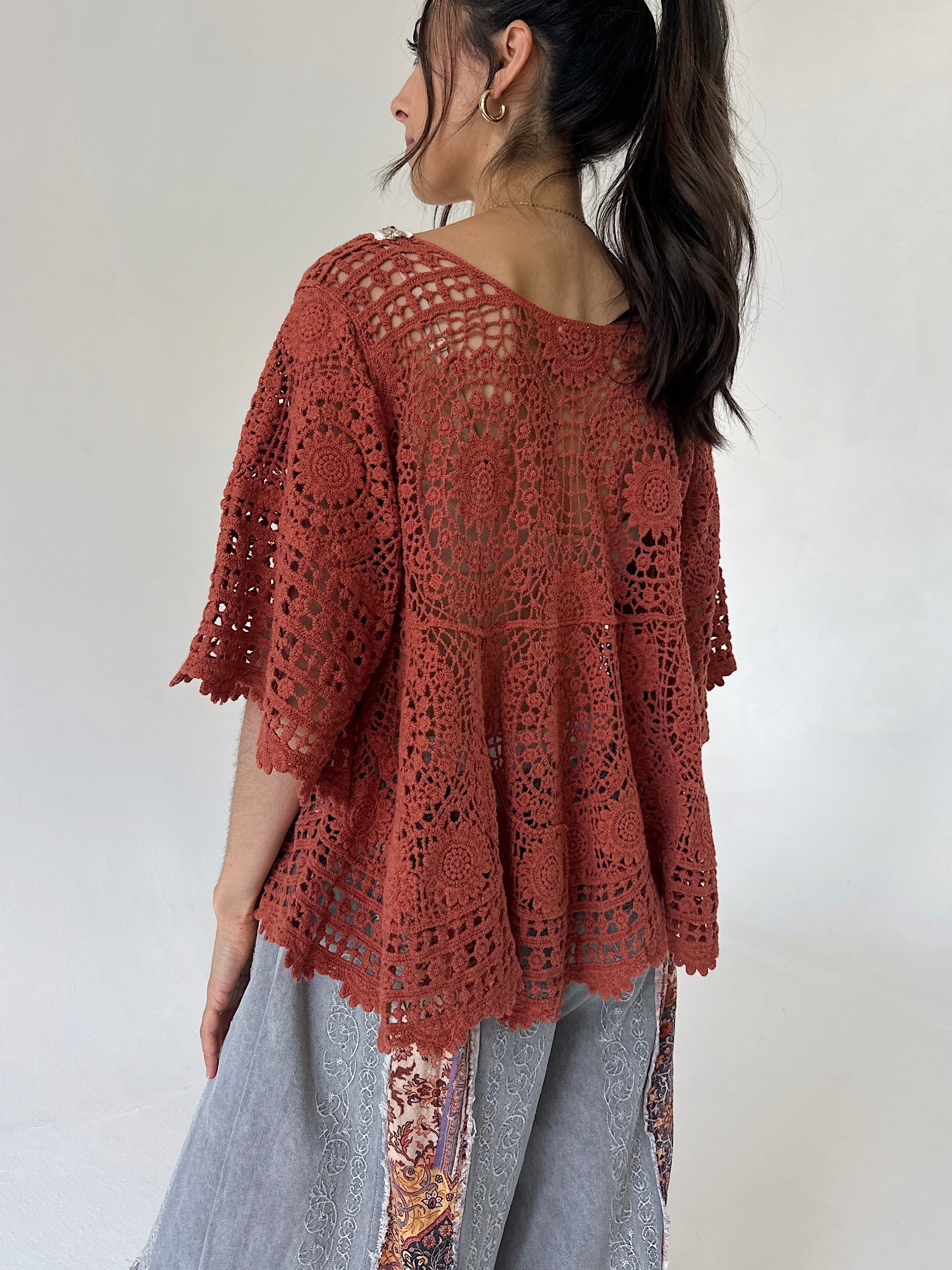 A woman shown from the back wearing a rust-colored, open-knit crochet style top over light gray patterned pants, set against a plain light gray background.