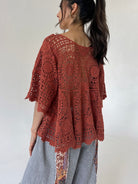 A woman shown from the back wearing a rust-colored, open-knit crochet style top over light gray patterned pants, set against a plain light gray background.