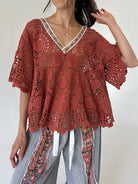 A woman wearing a rust-colored crochet lace top with a V-neck and short sleeves, paired with patterned gray pants, against a plain light background.