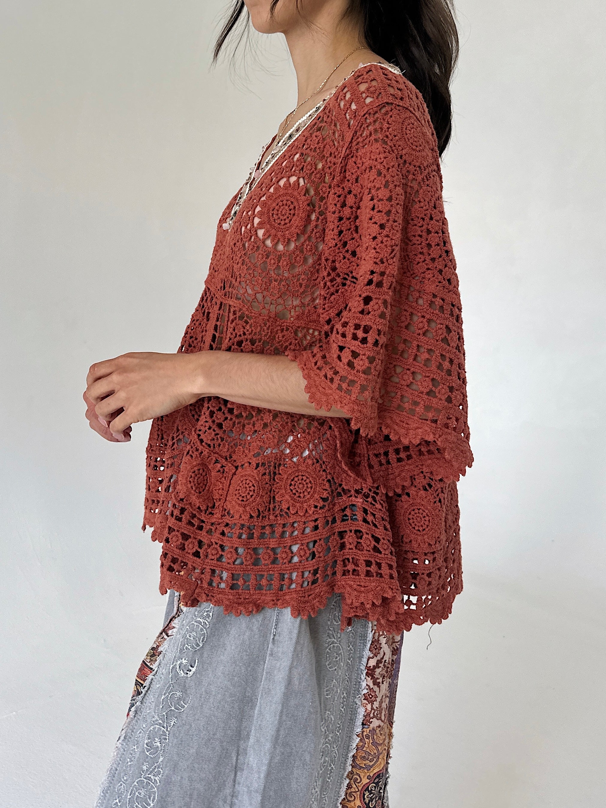 A woman wearing a rust-colored crochet poncho top with wide three-quarter sleeves and a scalloped hem, paired with light gray patterned pants, shown from a side profile against a plain light background.