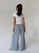 A woman viewed from behind wearing wide-leg gray pants with a white polka dot pattern, paired with a white t-shirt, standing against a plain light gray studio background.