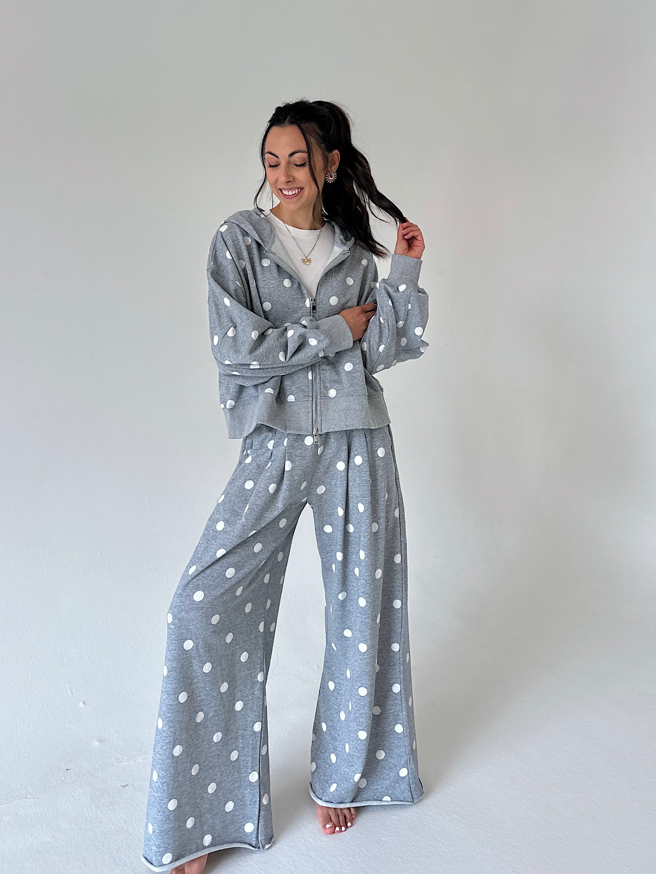A woman modeling a matching gray and white polka dot loungewear set, consisting of a zip-up hoodie and wide-leg pants, against a plain light gray studio background.